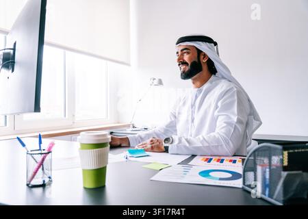 Young smiling arab man wearing traditional emirati kandura and ghutra, analyzing sales reports and digital data while working on a computer in a brigh Stock Photo