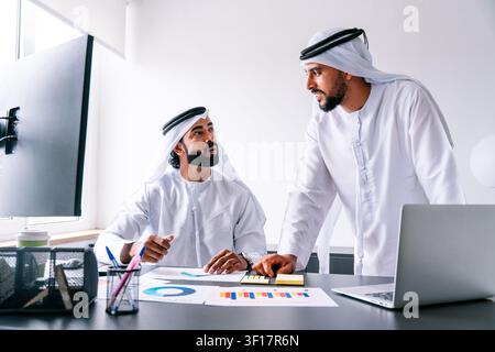 Two arab middle-eastern business men wearing traditional kandura discussing company reports and financial charts, reviewing data and making decisions Stock Photo