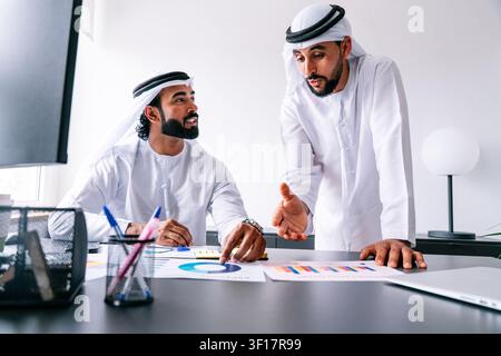 Two emirati businessmen are discussing business analytics and financial reports, pointing at data charts on the table while collaborating on a new str Stock Photo