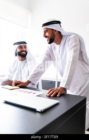 Two smiling adult arab men in traditional clothing collaborating in a modern office, standing and sitting while discussing business over documents and Stock Photo