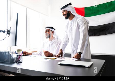 Two smiling arab businessmen wearing traditional kanduras are collaborating, reviewing data and discussing business strategies at a desk in a contempo Stock Photo