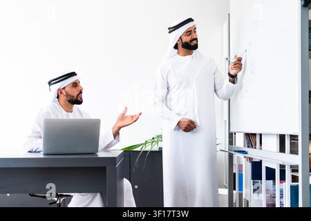 Two arab business colleagues collaborating in a modern office, with one man presenting ideas on a whiteboard while the other listens and comments from Stock Photo