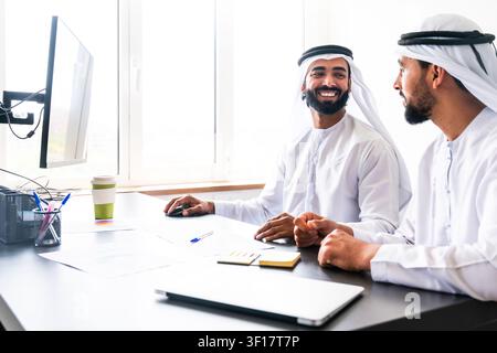 Two smiling arab businessmen wearing traditional emirati clothing are having a discussion and collaborating on a project in a modern office, one using Stock Photo