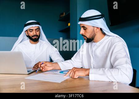 Two arab businessmen in traditional dress reviewing financial documents and interacting with a laptop, collaborating on strategic plans and analyzing Stock Photo