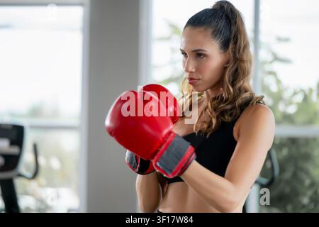 Portrait of a confident young woman wearing red boxing gloves in a gym, with a determined and focused gaze while practicing boxing and exercising. Sce Stock Photo