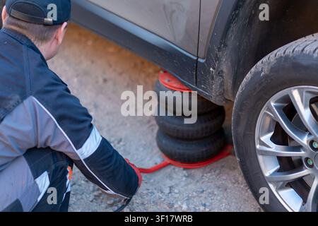 A mechanic is working under a vehicle lifted by a stack of tires in a gravel area. The mechanic wears a work suit and uses tools for repairs. The sett Stock Photo
