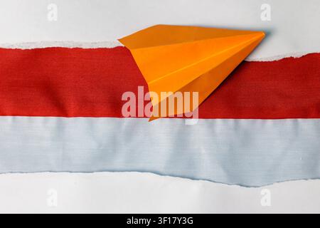 A creative top view composition featuring an orange paper airplane resting on a red and white fabric strip, framed by torn white paper borders. High Quality Photo Stock Photo