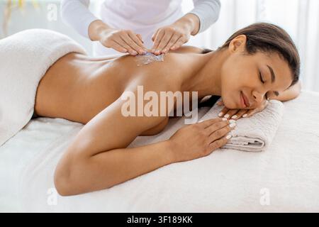 Therapist applying salt scrub to young black womans back at spa salon Stock Photo