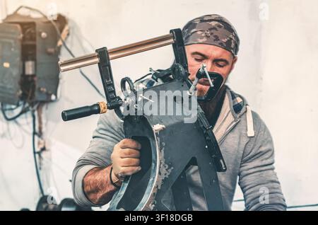 Metal frame in clamp in front of bearded man in bandana during repair in production workshop. Industrial background. Stock Photo