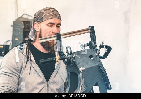 Metal frame in clamp in front of bearded man in bandana during repair in production workshop. Industrial background. Stock Photo