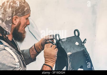 Bearded man in bandana tightens bolt into metal frame part during repair. Side view of craftsman in workshop against white wall background. Stock Photo