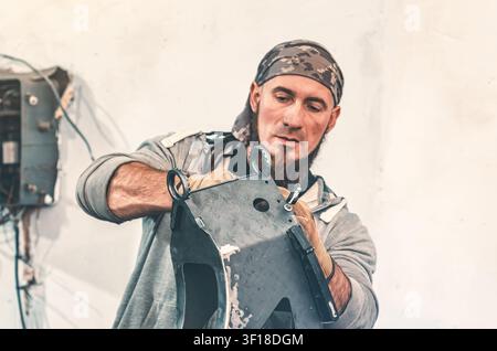 Bearded man in bandana holding metal frame part while working on repair. Portrait of craftsman in workshop against white wall. Stock Photo