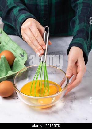 Person in plaid shirt whisking eggs with green whisk in glass bowl on marble table close-up Stock Photo