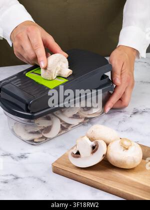 Woman's hands slicing mushrooms on a handheld vegetable slicer with green blade on marble table close-up Stock Photo