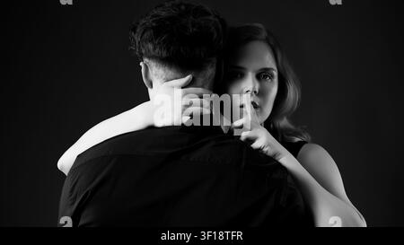 Woman embracing man from behind, looking at camera and making hush gesture Stock Photo