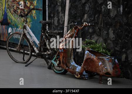 A rusted bicycle and weathered scooter stand against a graffiti-marked wall in Amsterdam, showing peeling paint, corroded metal and weeds on paving. Stock Photo