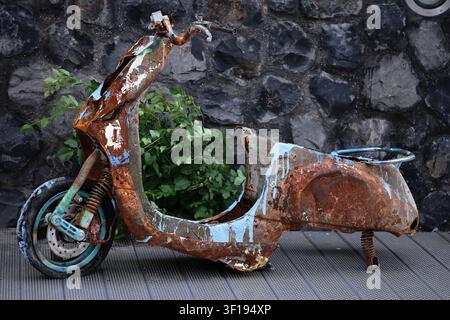Close view of a heavily rusted scooter beside a dark stone wall in Amsterdam, with flaking paint, bent metal and green weeds on paving at its base. Stock Photo