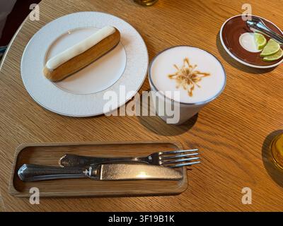 Fresh eclair with vanilla glaze is served beside creamy latte on wooden table in modern cafe with elegant table setting. French pastry, coffee break Stock Photo