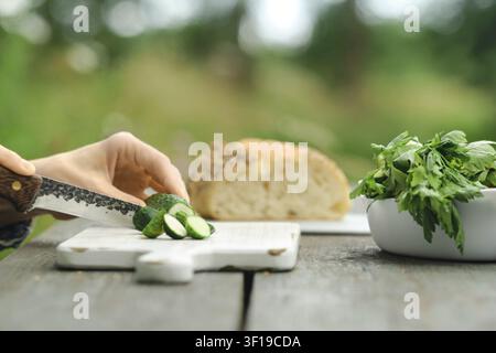 Person slices cucumbers on wooden cutting board. Fresh bread sits in background. Green herbs are nearby. Scene takes place outdoors in daylight with b Stock Photo