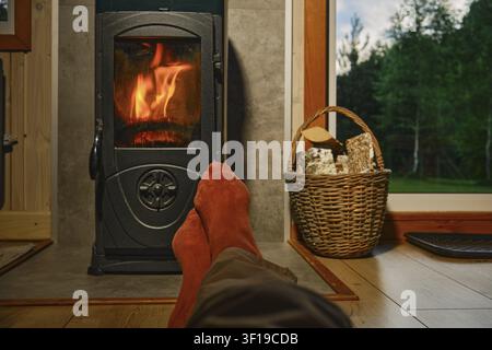 Feet in red socks are resting in front of wood stove in wooden cabin. Flames are visible, creating warmth. A basket with firewood sits nearby. Cabin i Stock Photo