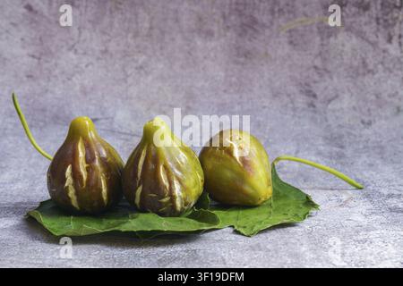 Ripe brown and green figs sitting on green fig leaves, against a rustic, textured grey background Stock Photo