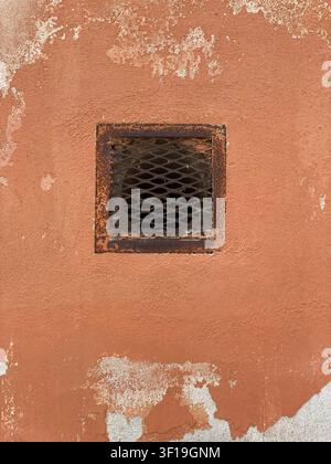 Cremona, Italy June 19th 2026 Weathered terracotta colored wall featuring an old square rusty metal grate, revealing layers of peeling paint and cemen Stock Photo