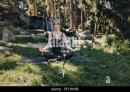Woman records yoga class on smartphone mounted on tripod in front of waterfall. Online yoga instructor, virtual wellness coaching, meditation app Stock Photo