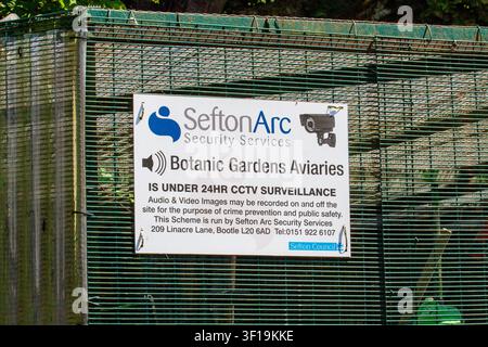 A security sign on an aviary cage reads "Sefton Arc Security Services Botanic Gardens Aviaries IS UNDER 24HR CCTV SURVEILLANCE" in public park setting. Stock Photo