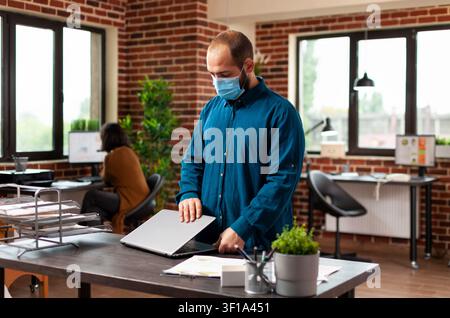 Masked executive opening laptop before startup meeting, bookkeeper planning business meeting, showing financial data, focused on masked executive. Stock Photo