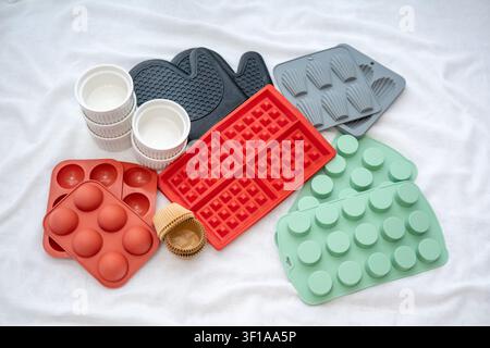 The red waffle silicone molds and the green round pans with the ceramic ramekins and the black oven mitts lie on the white cloth on the table. Stock Photo