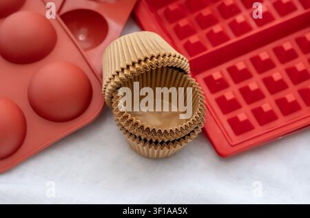 The brown paper cupcake liners, the red silicone waffle molds, and the round half-spheres lie on the white linen fabric on the flat table. Stock Photo