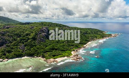 Aerial view of a coastline with crystal clear water, lush green forest,and waves breaking on the shore. La Digue, Seychelles. Stock Photo