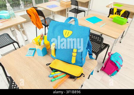 Blue school backpack with yellow straps and pencil case on a wooden desk in an empty classroom setting Stock Photo