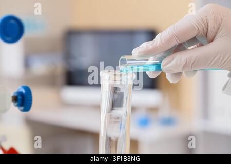 Scientist in white glove pouring blue liquid from a test tube into a beaker in a laboratory setting Stock Photo