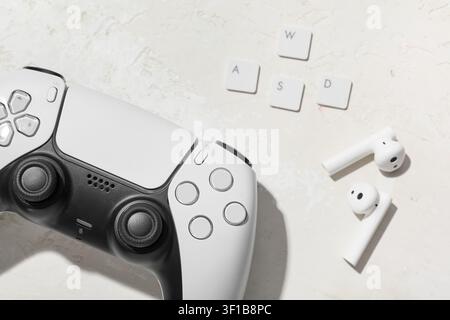 White video game controller with wireless earbuds and keyboard keys on a textured background, gaming setup Stock Photo