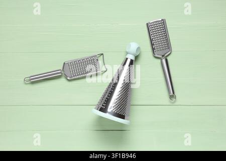 Three stainless steel kitchen graters with different shapes and handles on a light green wooden background, the graters include a cone-shaped grater w Stock Photo