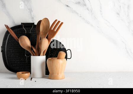 Wooden kitchen utensils in a white holder with a grill pan and mortar and pestle on a textured white surface Stock Photo