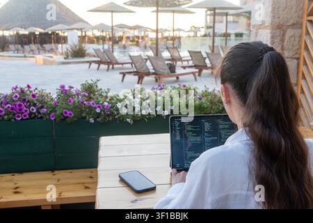 Woman with dark hair in a ponytail sits at a wooden table programming on a tablet computer at an outdoor resort cafe. Stock Photo
