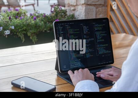 Close-up over the shoulder of a woman typing code on a tablet computer keyboard at a wooden table in an outdoor resort cafe. Stock Photo