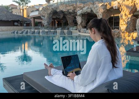 Brunette woman in white apparel sits on a poolside sunbed working with a tablet computer displaying analytics charts against artificial rock formation Stock Photo