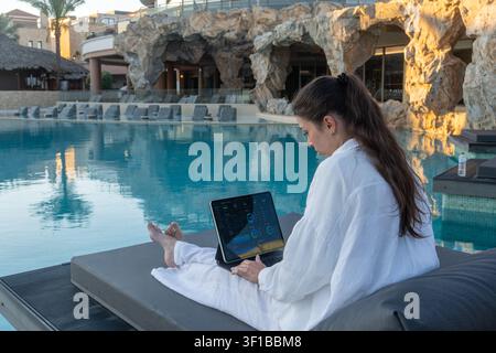 Brunette woman in white apparel sits on a poolside sunbed working with a tablet computer displaying analytics charts against artificial rock formation Stock Photo