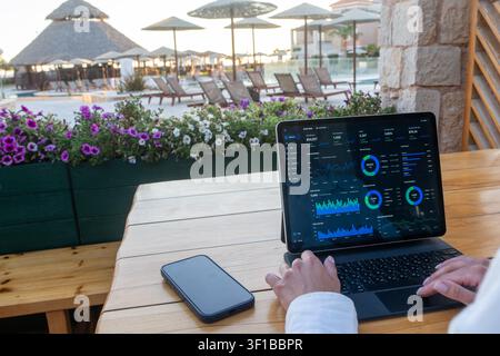 Tablet displaying financial data charts sits on a wooden table with a smartphone at an outdoor resort cafe while hands type. Stock Photo