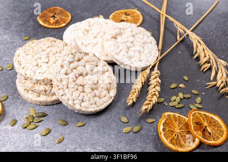 Several round rice cakes with pumpkin seeds, dried orange slices, and wheat ears scattered on a dark gray surface, representing healthy snack options Stock Photo