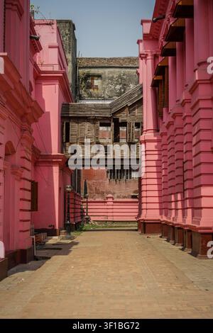 Two pink palace facades with multiple columns flank a brick walkway, leading towards a rustic wooden bridge connecting two older, weathered buildings. Stock Photo