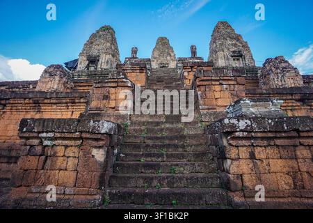 A wide, steep stone staircase covered in dark green moss and small plants leads upwards towards three large rust-colored brick towers, with two smalle Stock Photo