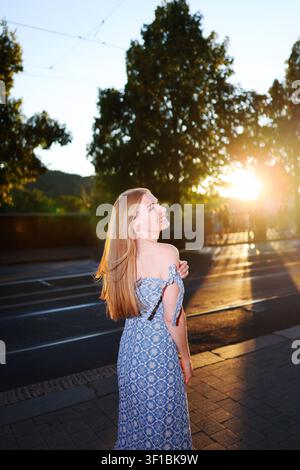 Young woman looking back with a smile while strolling through a European city at sunset, capturing freedom and summer vacation mood. Stock Photo