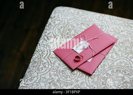 Pink wedding invitation envelopes with gold rings and floral tag lie on patterned tablecloth Stock Photo