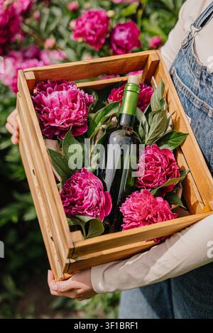 Woman Holding Homemade White Wine Bottle Surrounded by Garden Flowers. Small-scale winemaking, entrepreneurship, local production and artisan craftsmanship concept. Stock Photo