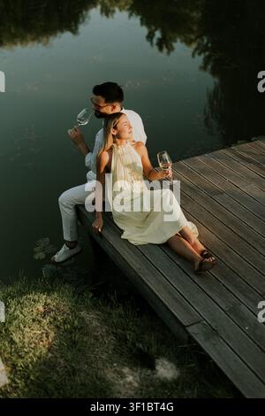 A man and a woman relax together on a wooden dock. They hold glasses filled with drinks as they sit by the water. The sun is setting behind them, cast Stock Photo