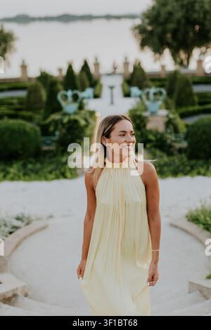 A woman walks down stone steps in a garden filled with plants. She smiles and wears a light dress. The sun sets behind the water, creating a soft glow Stock Photo